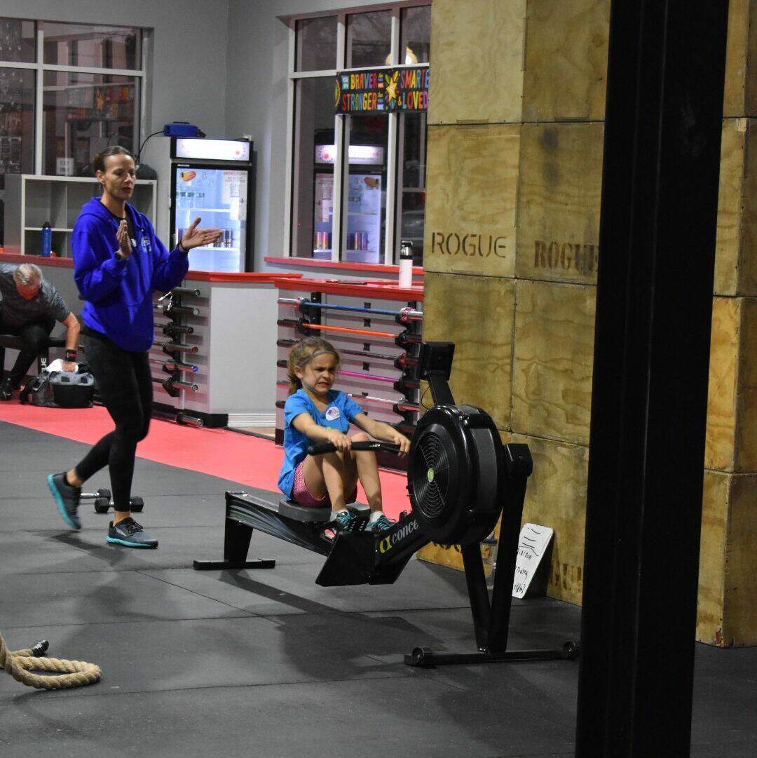 A woman in a blue sweatshirt stands next to a girl on a rowing machine in a gym