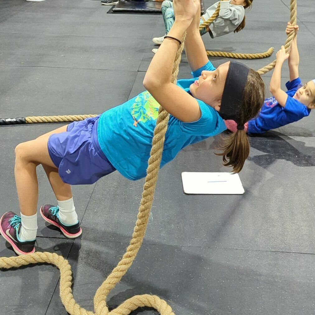 A girl in a blue shirt is climbing a rope in a gym