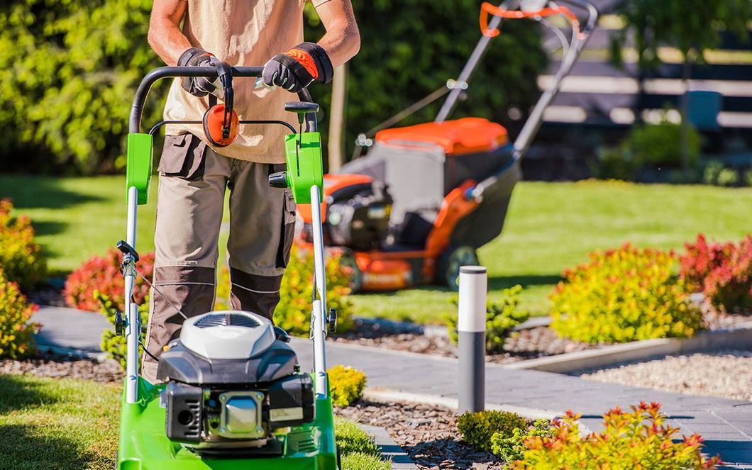 A person wearing work clothes and gloves pushes a green lawnmower in a landscaped garden with a second mower nearby.