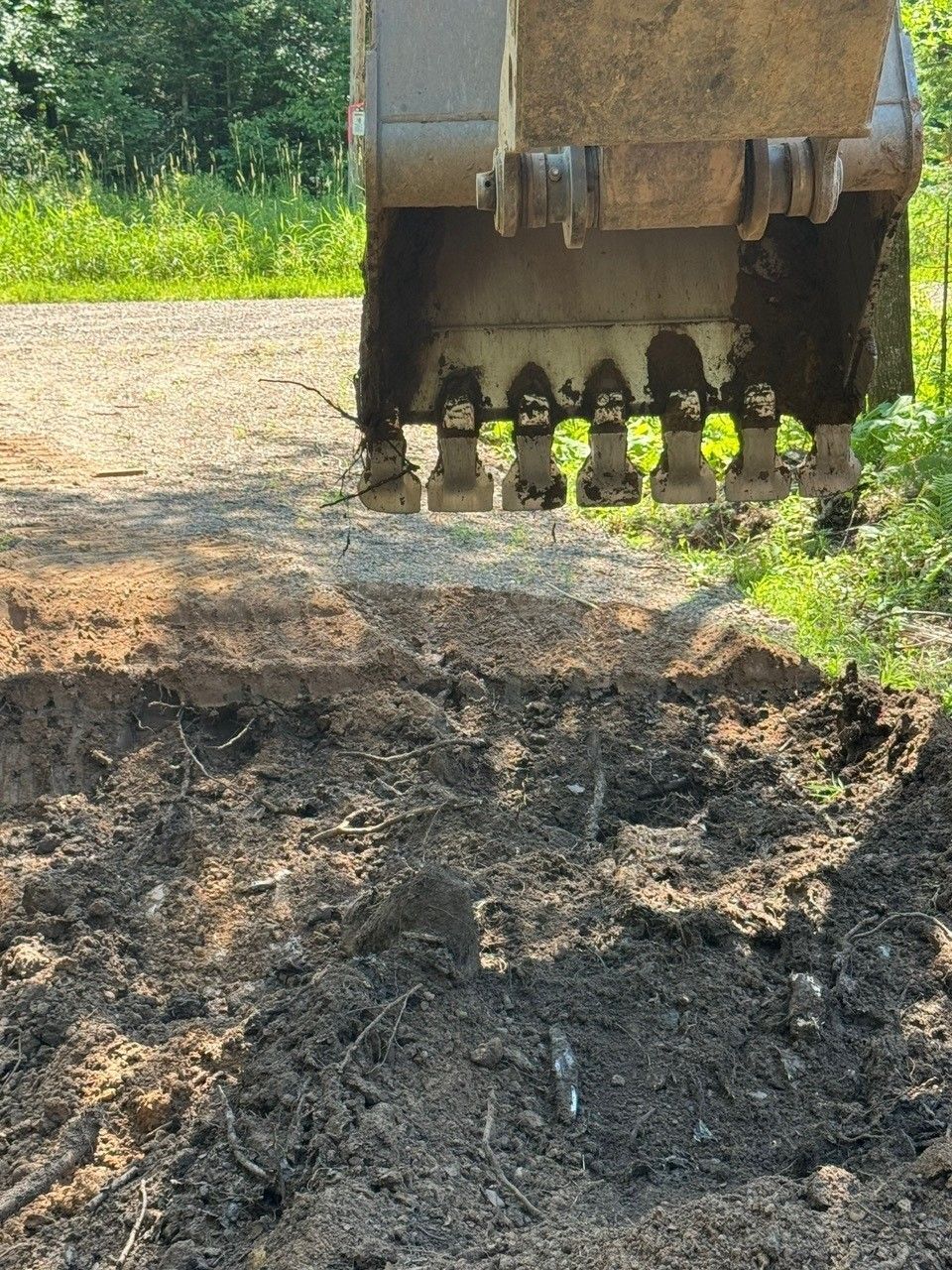 Excavator bucket digging into the ground on a dirt road, with a grassy tree line in the background.