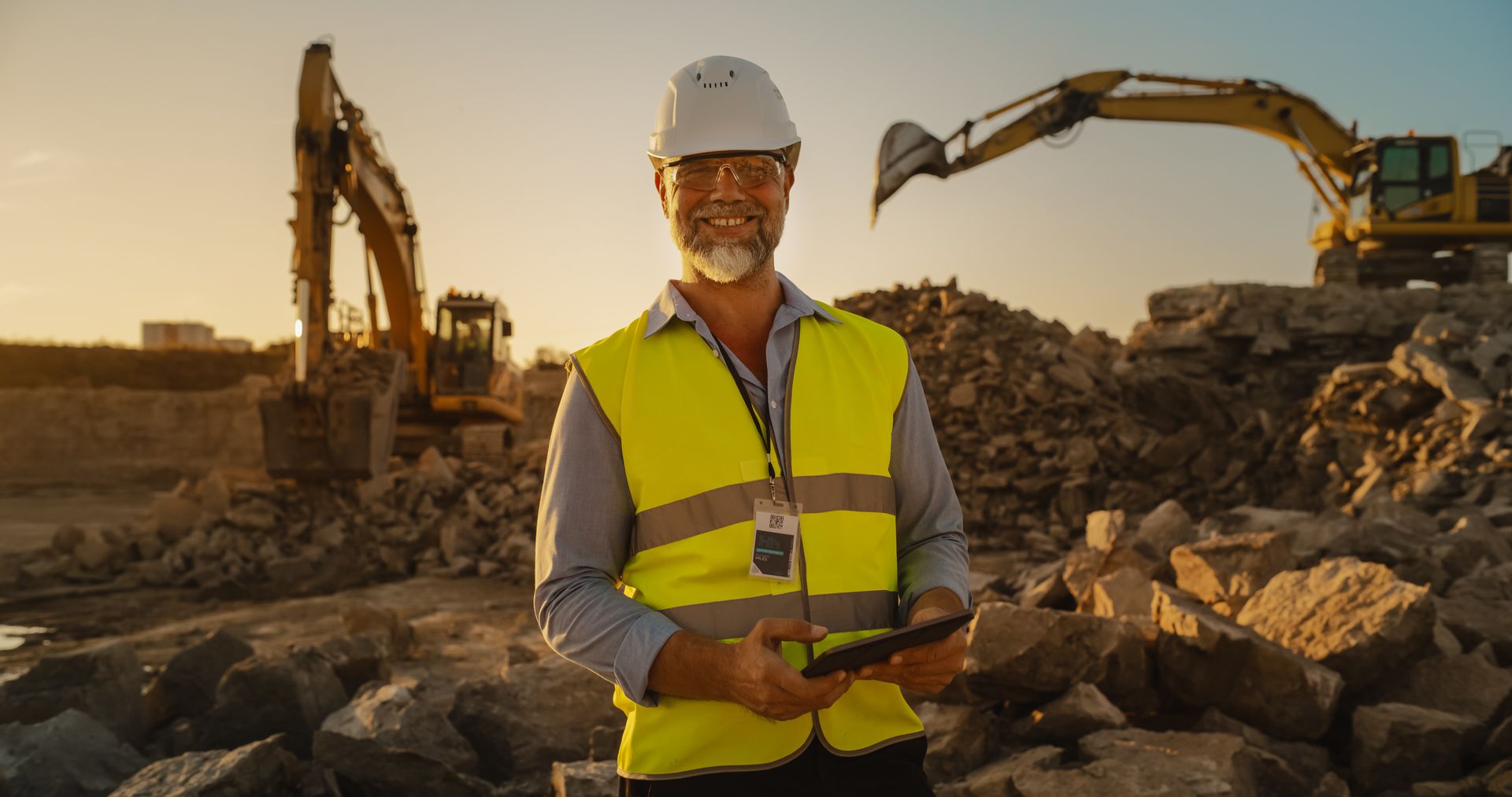 Smiling worker in hard hat and vest holds a tablet at a rocky excavation site with two excavators.