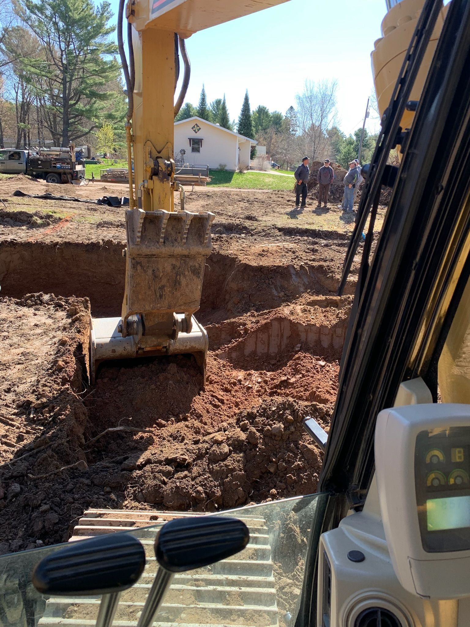 Excavator digging a foundation trench; a house and people are in the background. Sunny outdoor setting.