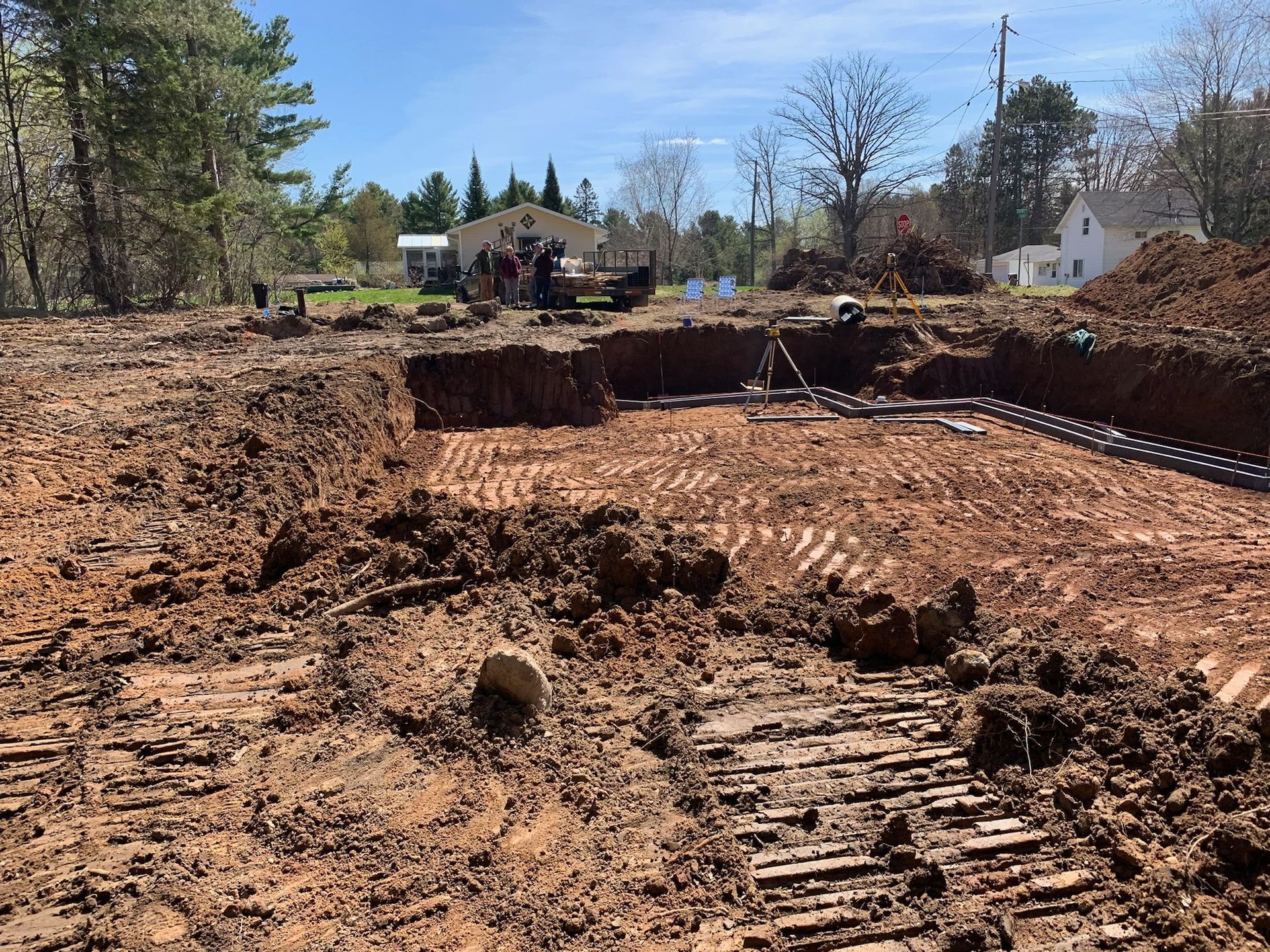 Construction site, freshly dug foundation pit with dirt mounds and equipment, houses in background.