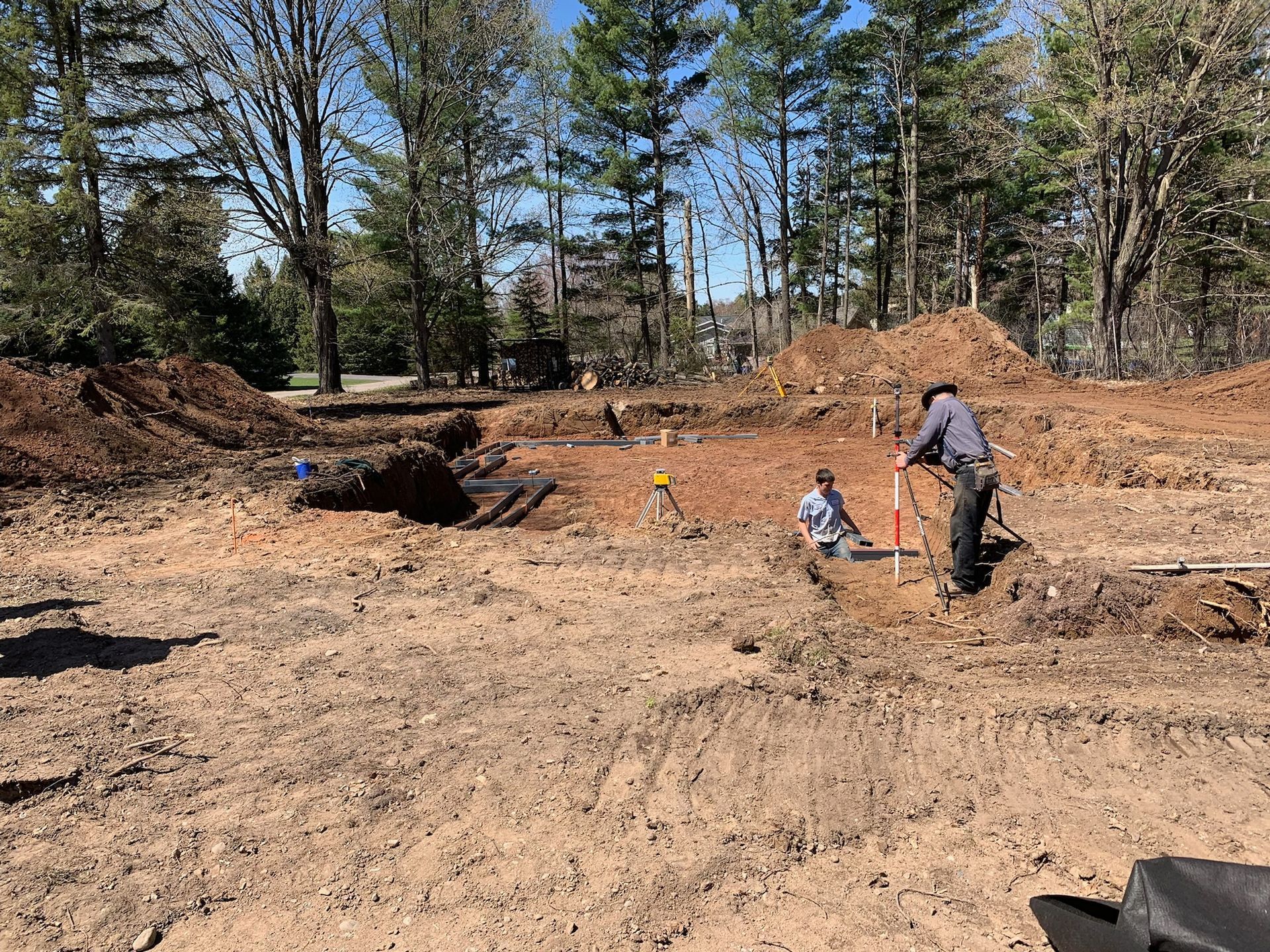 Construction site with two workers surveying the land. Brown soil, trees in the background, blue sky.