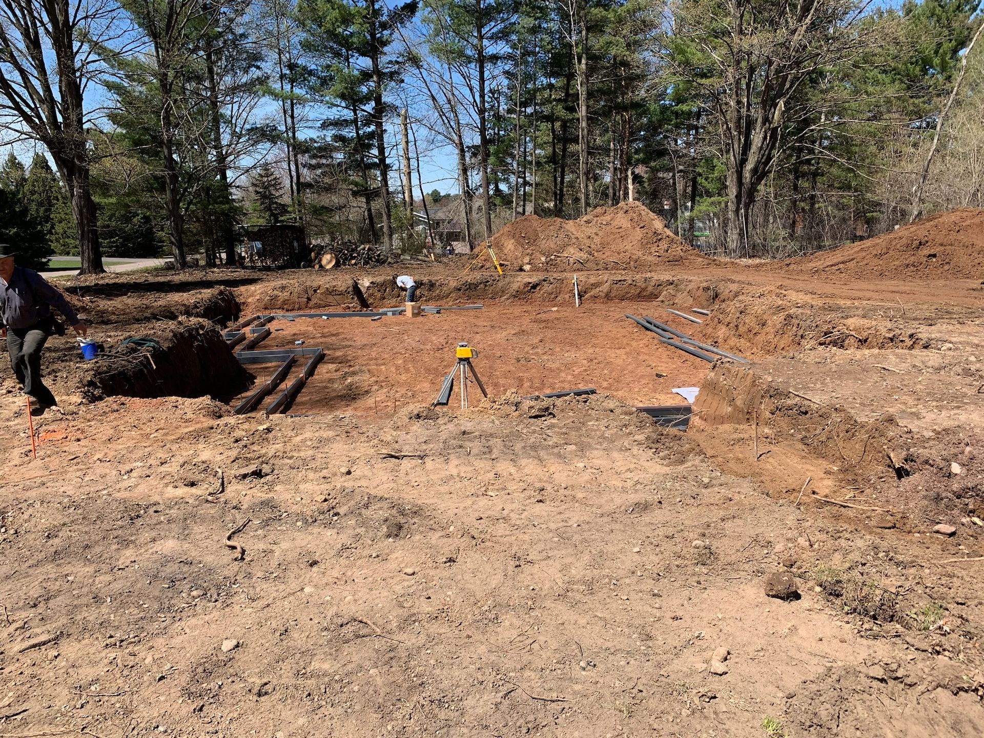 Construction site with trenches, dirt mounds, and workers; sunny, outdoor setting.