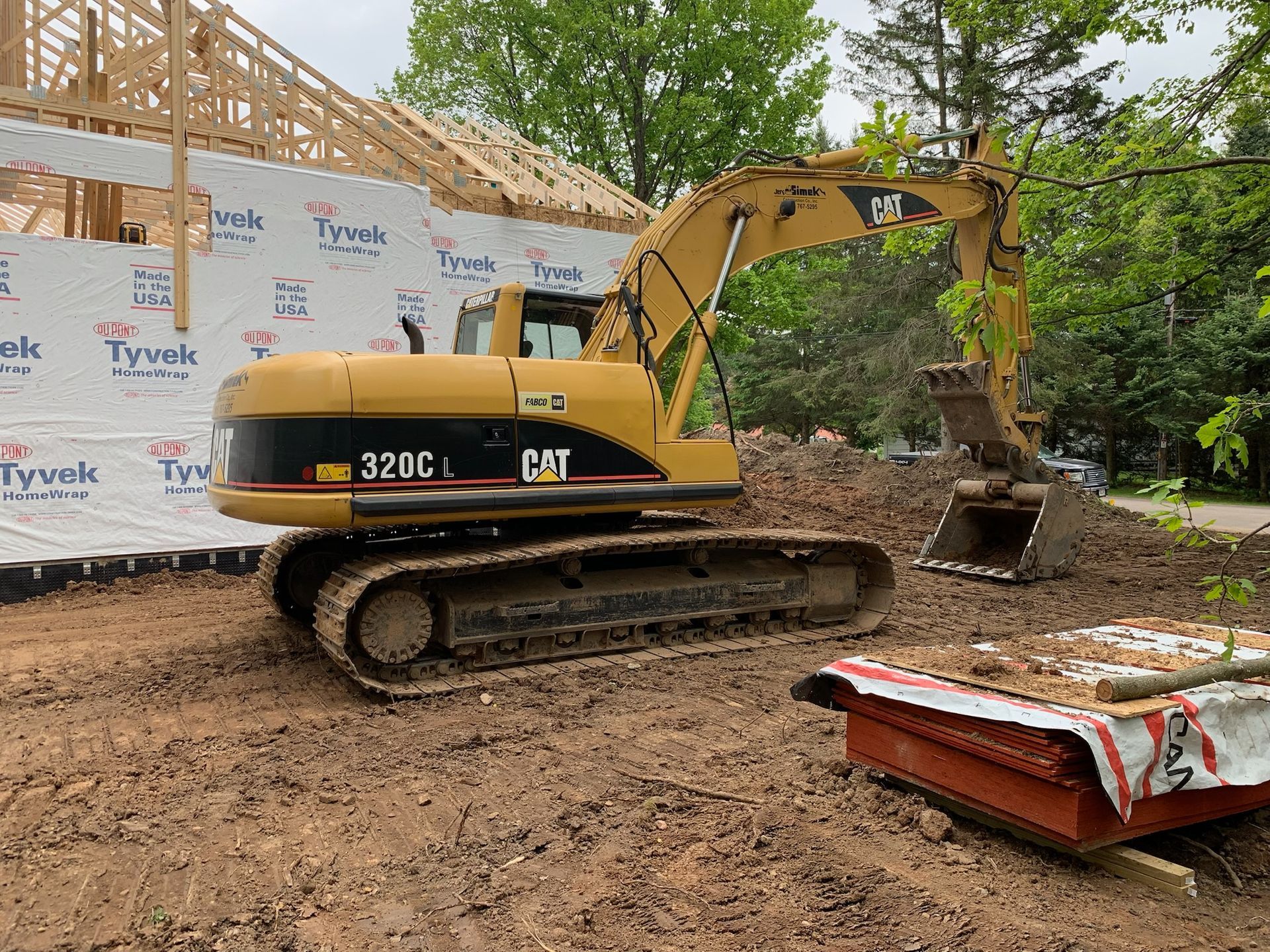Yellow Caterpillar 320C excavator at a construction site, digging in dirt next to a house under construction.