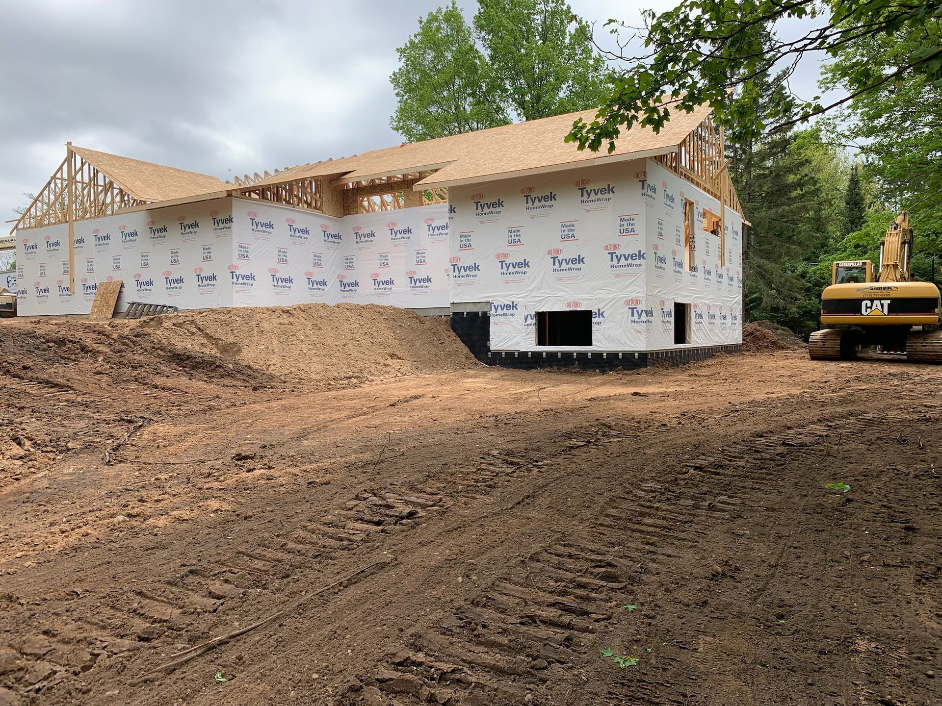 Construction of a building, walls wrapped in Tyvek, unfinished roof, excavator nearby on dirt.