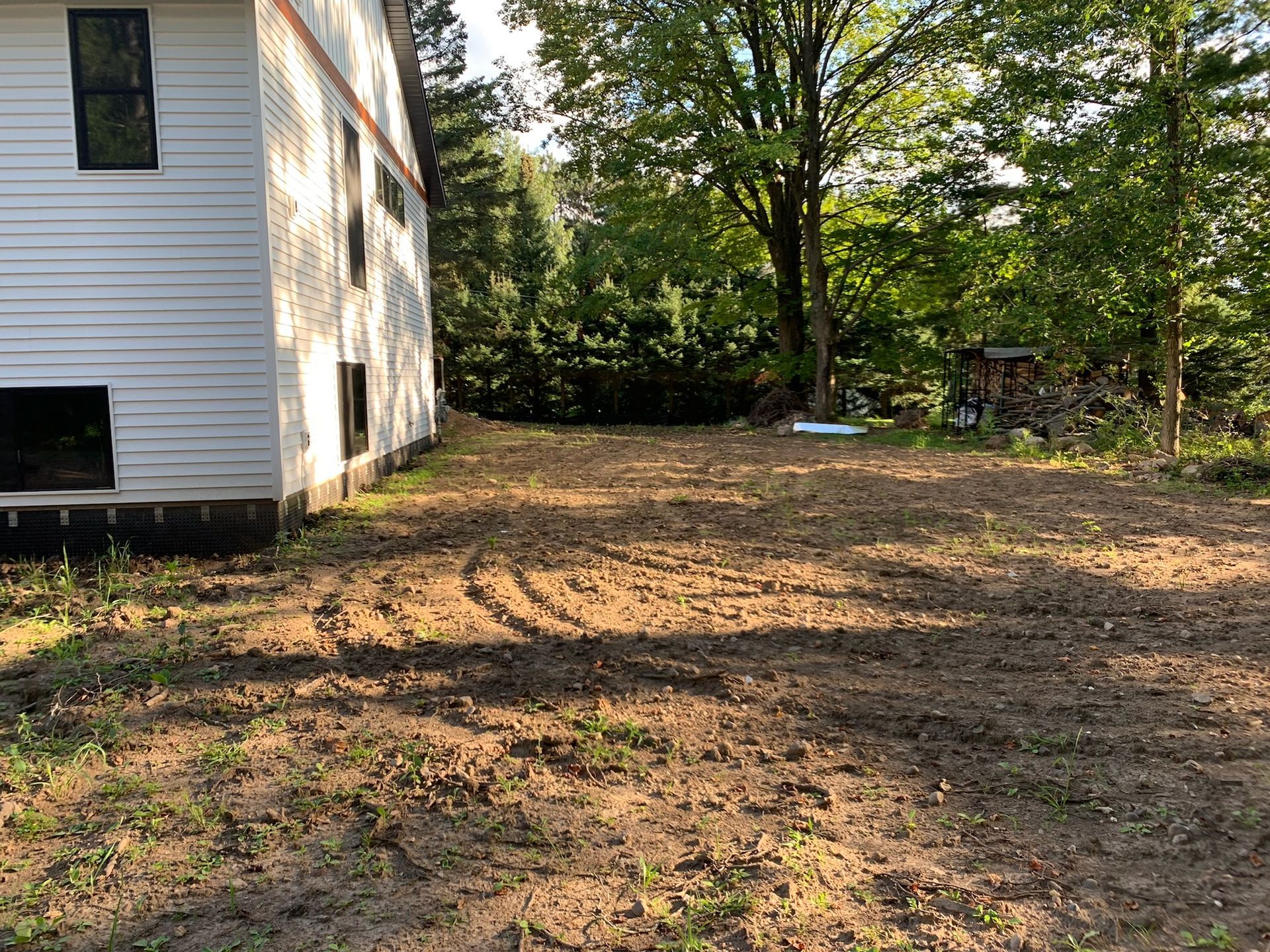 Cleared backyard with dirt, near a house with white siding and trees in the background.