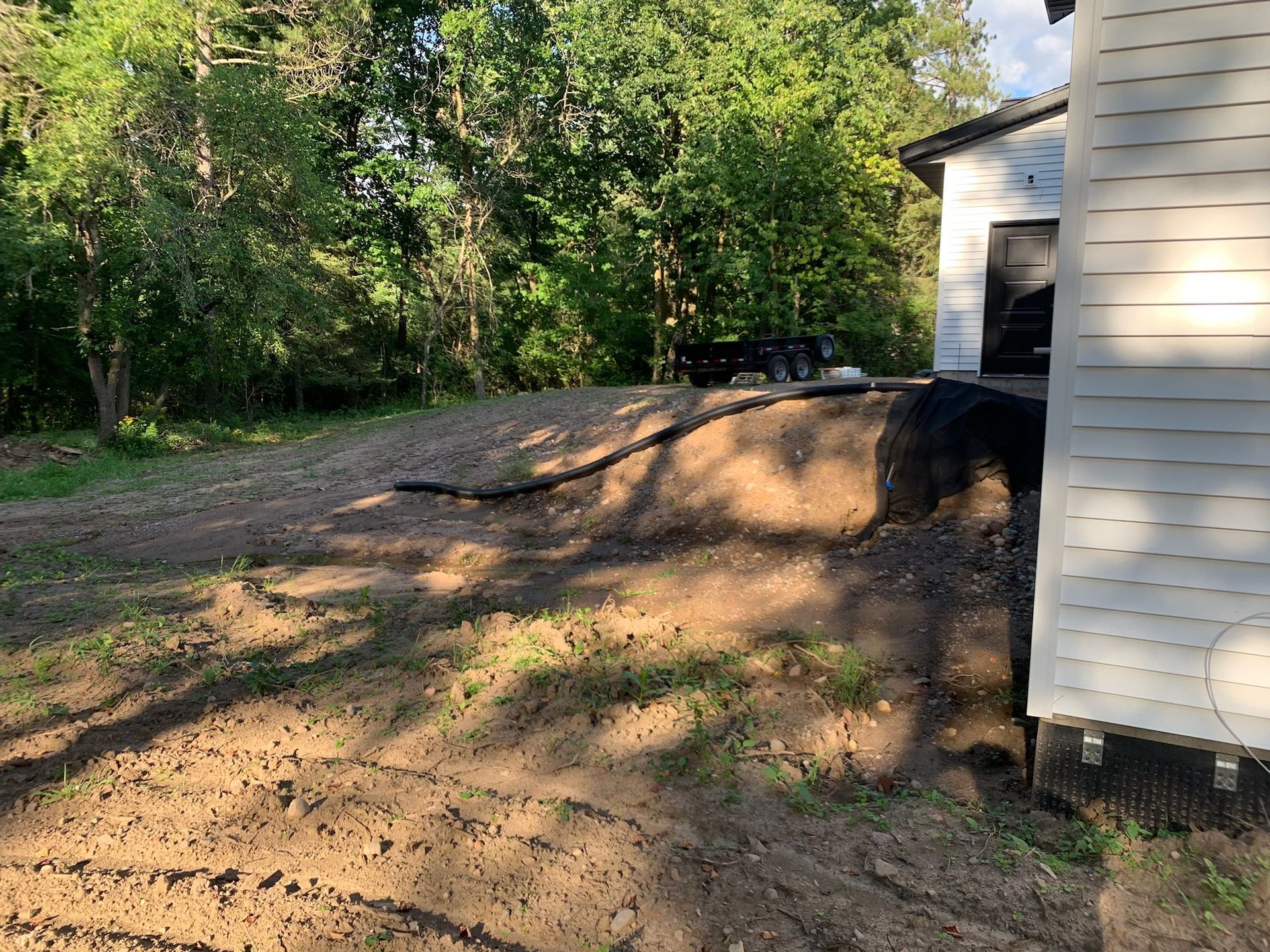 Dirt and gravel landscaping near a garage, with a black pipe and trees in the background.