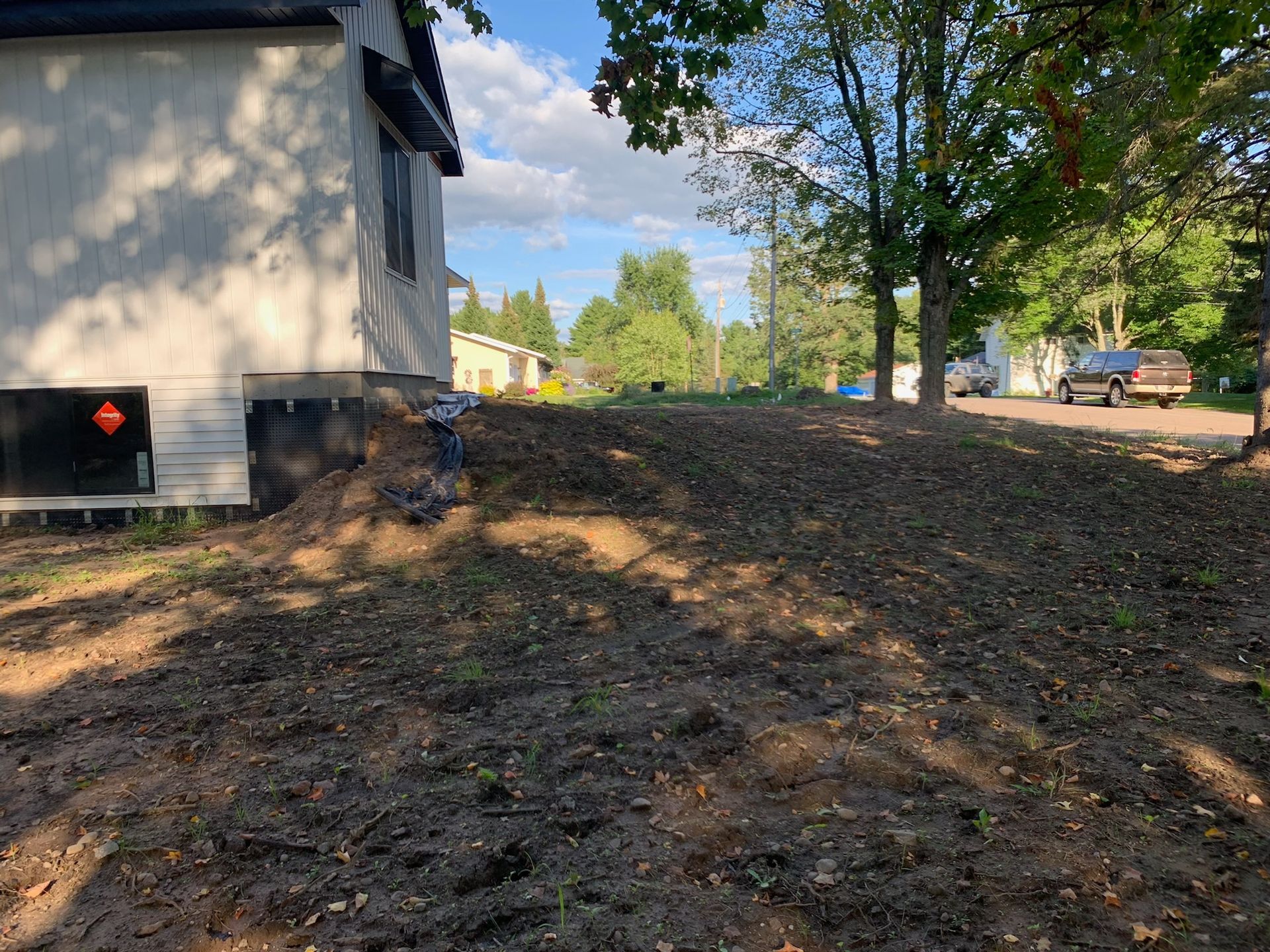 House next to a dirt hill, likely a newly landscaped area. Trees and a car are visible in the background.