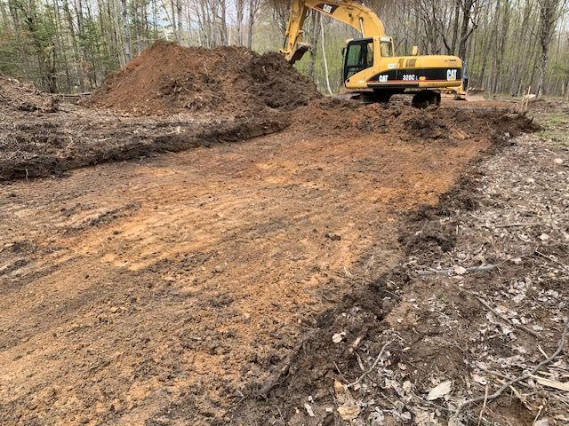 Yellow excavator digging in a dirt area, moving soil; forest background.