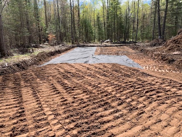 Excavated foundation site with dark soil, fabric, and tire tracks, surrounded by trees.