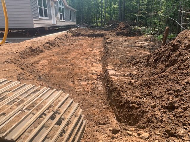 Excavated trenches in dirt for foundation next to a light-colored house in a wooded area.