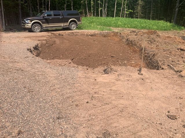 Brown truck beside a dirt clearing. Excavated soil piled around the perimeter, trees in background.