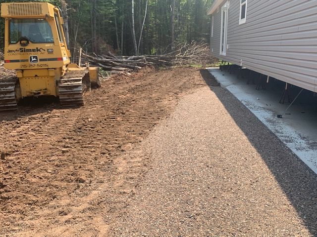 A yellow bulldozer and gravel pathway next to a modular home under construction in a wooded area.