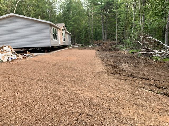 Gravel driveway in front of a gray house, with forest in the background.