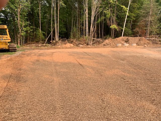 Gravel lot cleared of trees; a yellow bulldozer at left, with woods in the background.