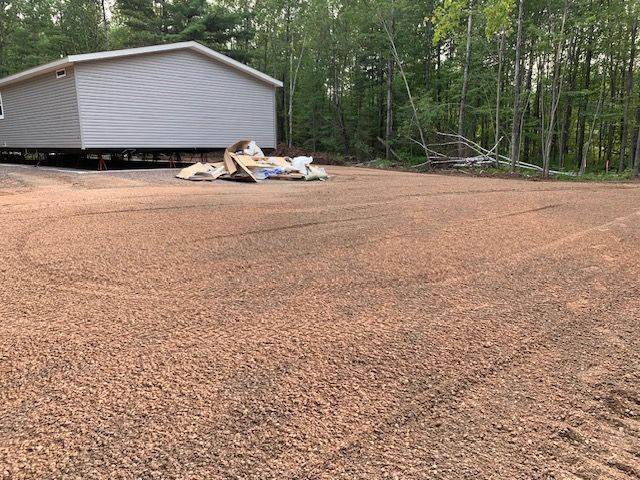A gravel driveway in front of a light gray house next to a wooded area. Debris on the ground.