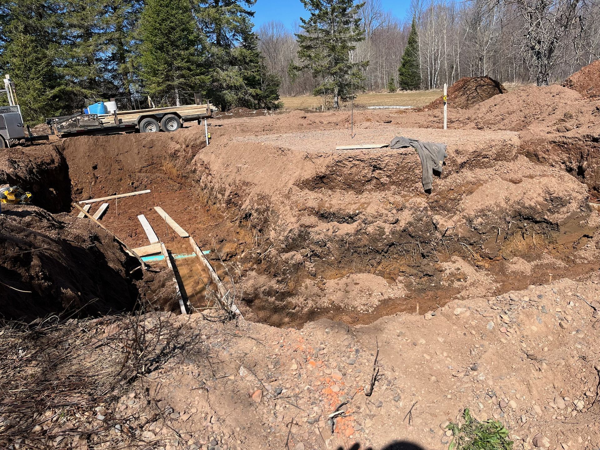 Excavation for a circular structure in progress; earth walls, wooden supports, trailer, trees in background.