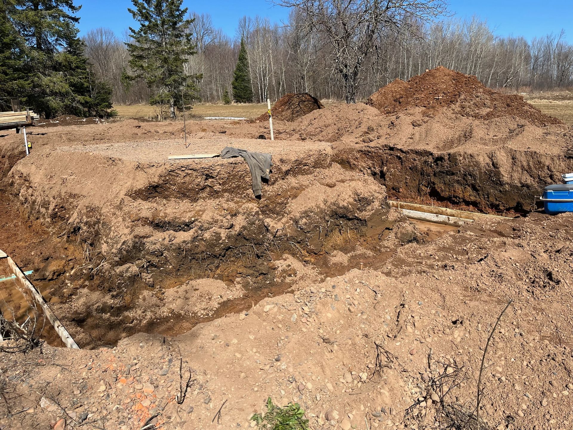 Dirt excavation for a foundation, showing soil layers, pipes, and a blue container outdoors on a sunny day.