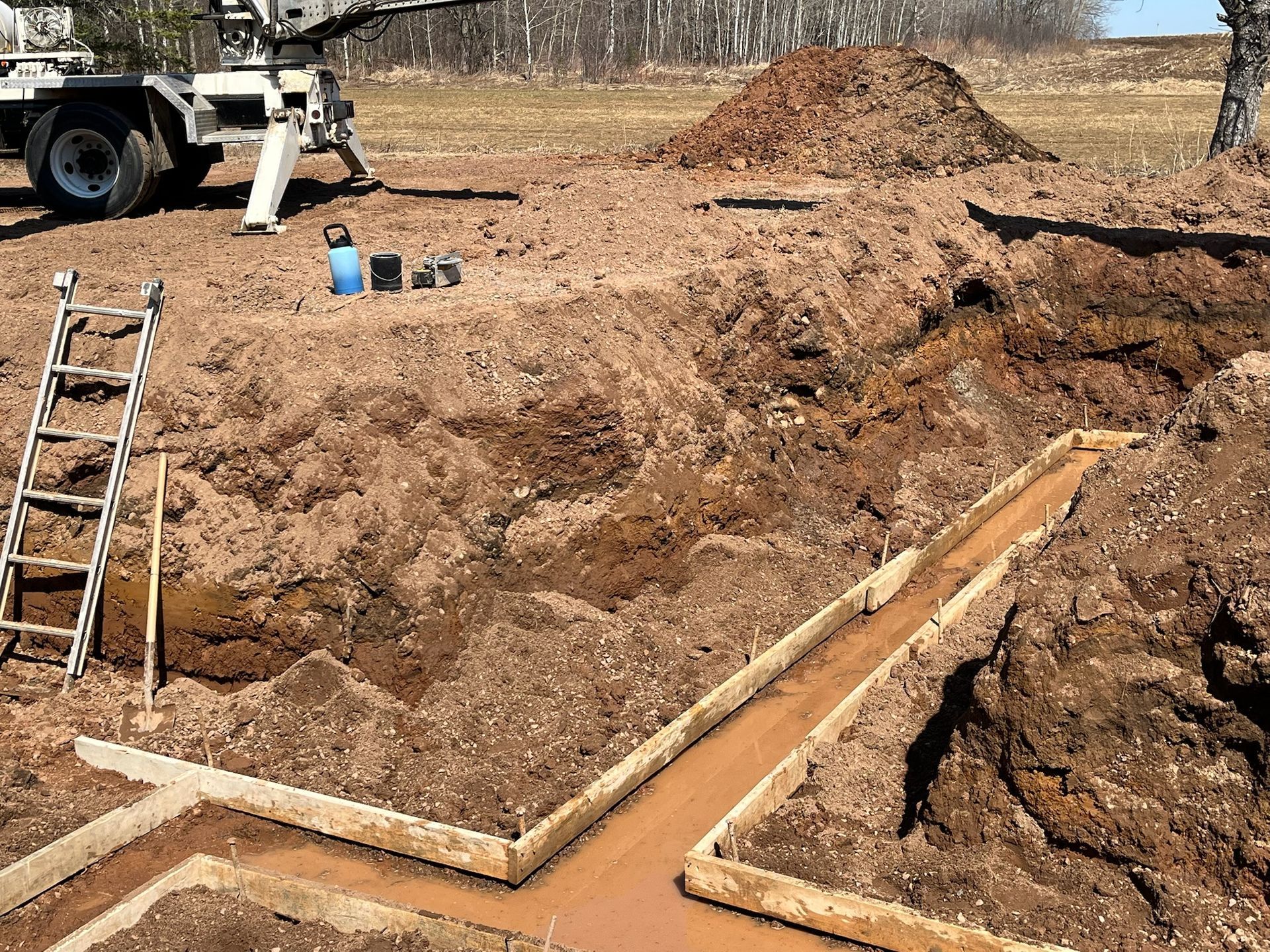 Construction site with trenches dug for foundation forms, dirt pile, ladder, and truck.