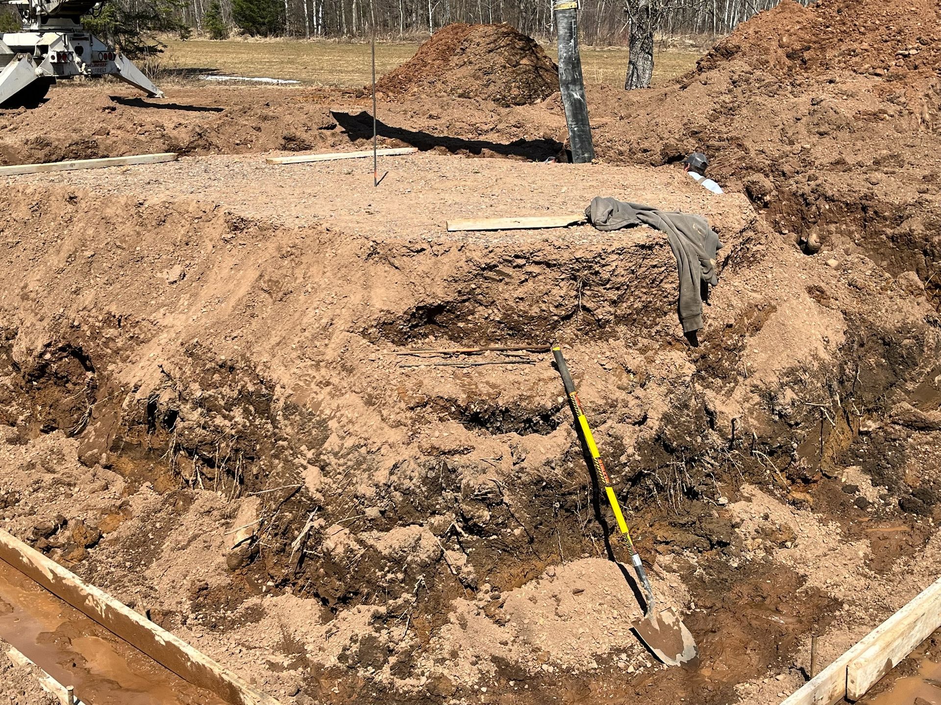 Excavated soil layers in construction site, with a shovel, wooden forms, and piles of dirt.