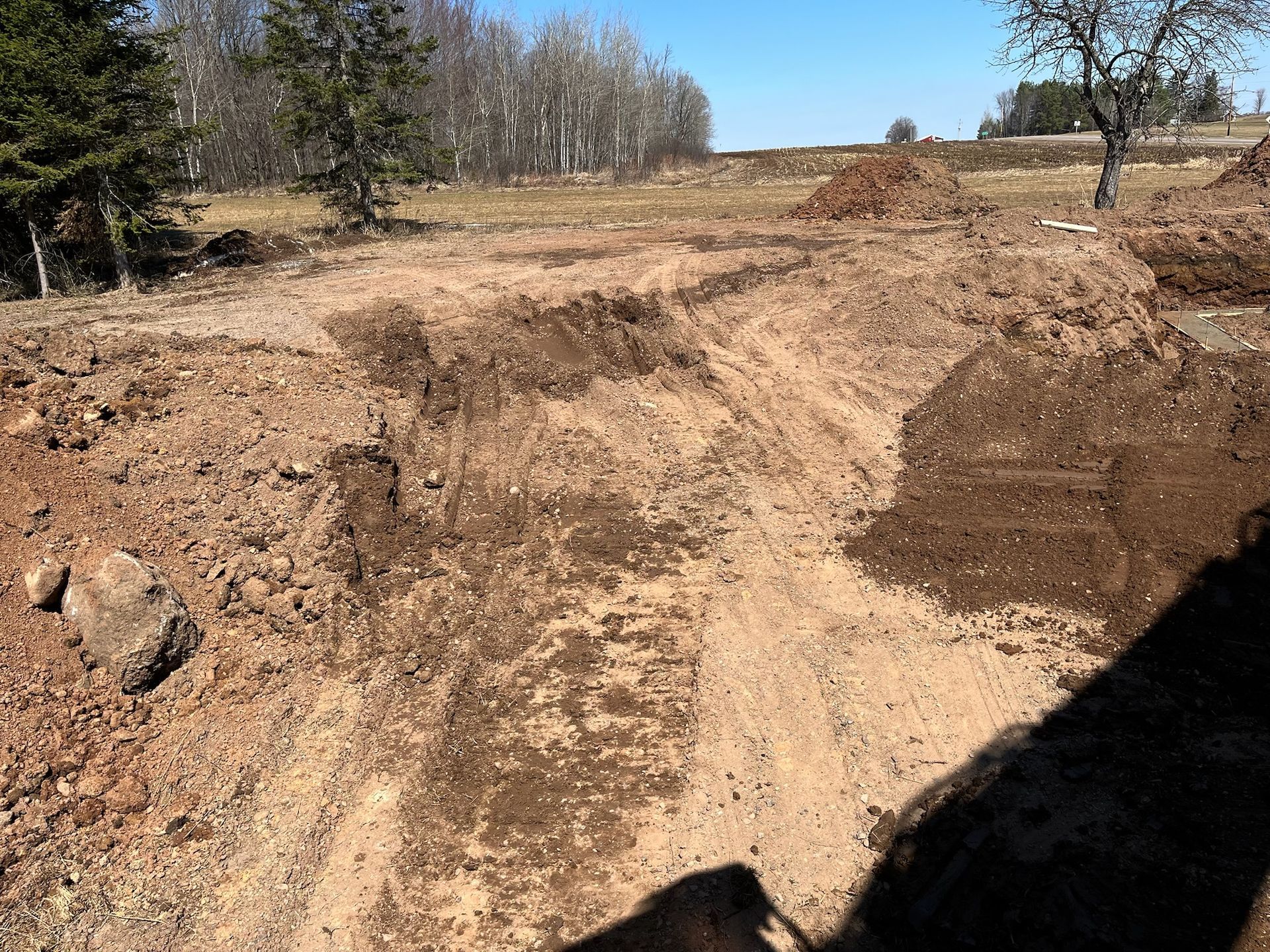 Dirt construction site with piles of excavated earth, a trench, and trees in the background under a blue sky.