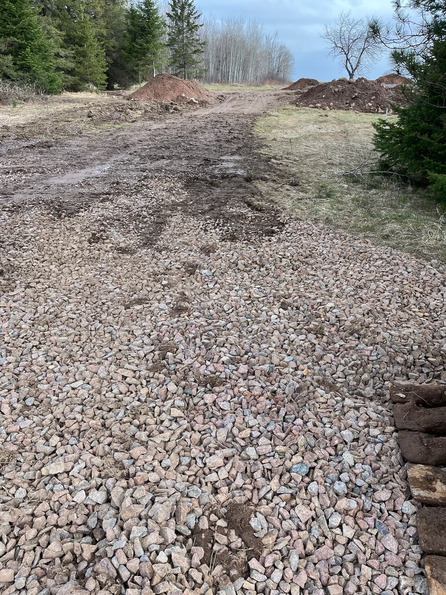 Gravel driveway leading uphill towards trees and a cloudy sky. Wet and muddy areas visible.