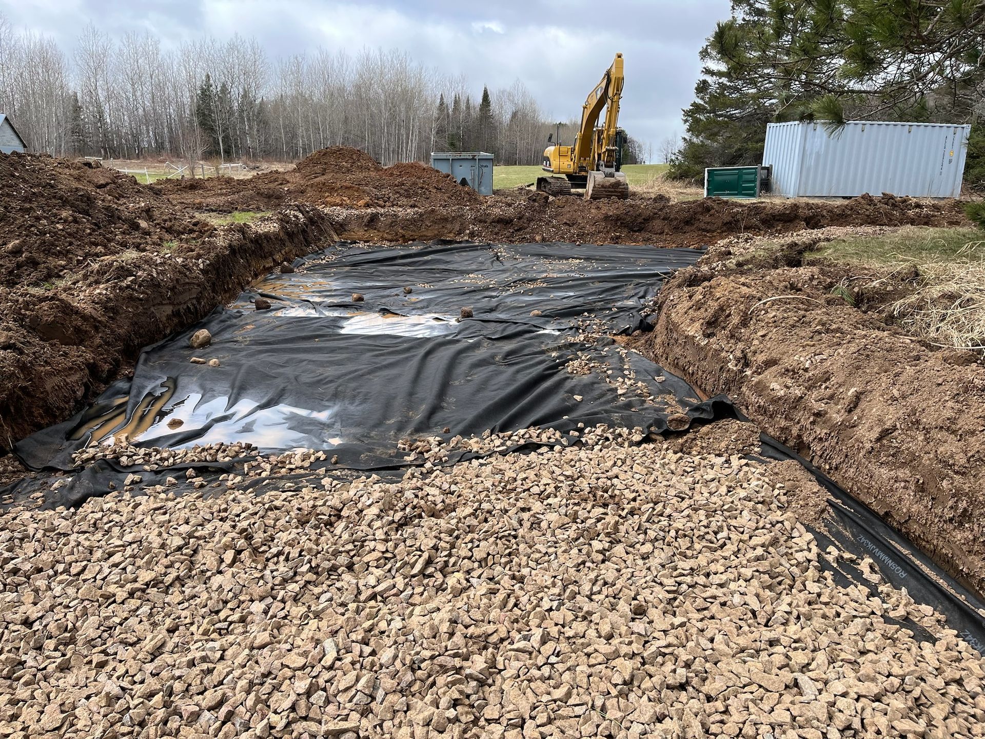 Construction site with excavated foundation covered by black tarp, gravel base, and an excavator in the background.