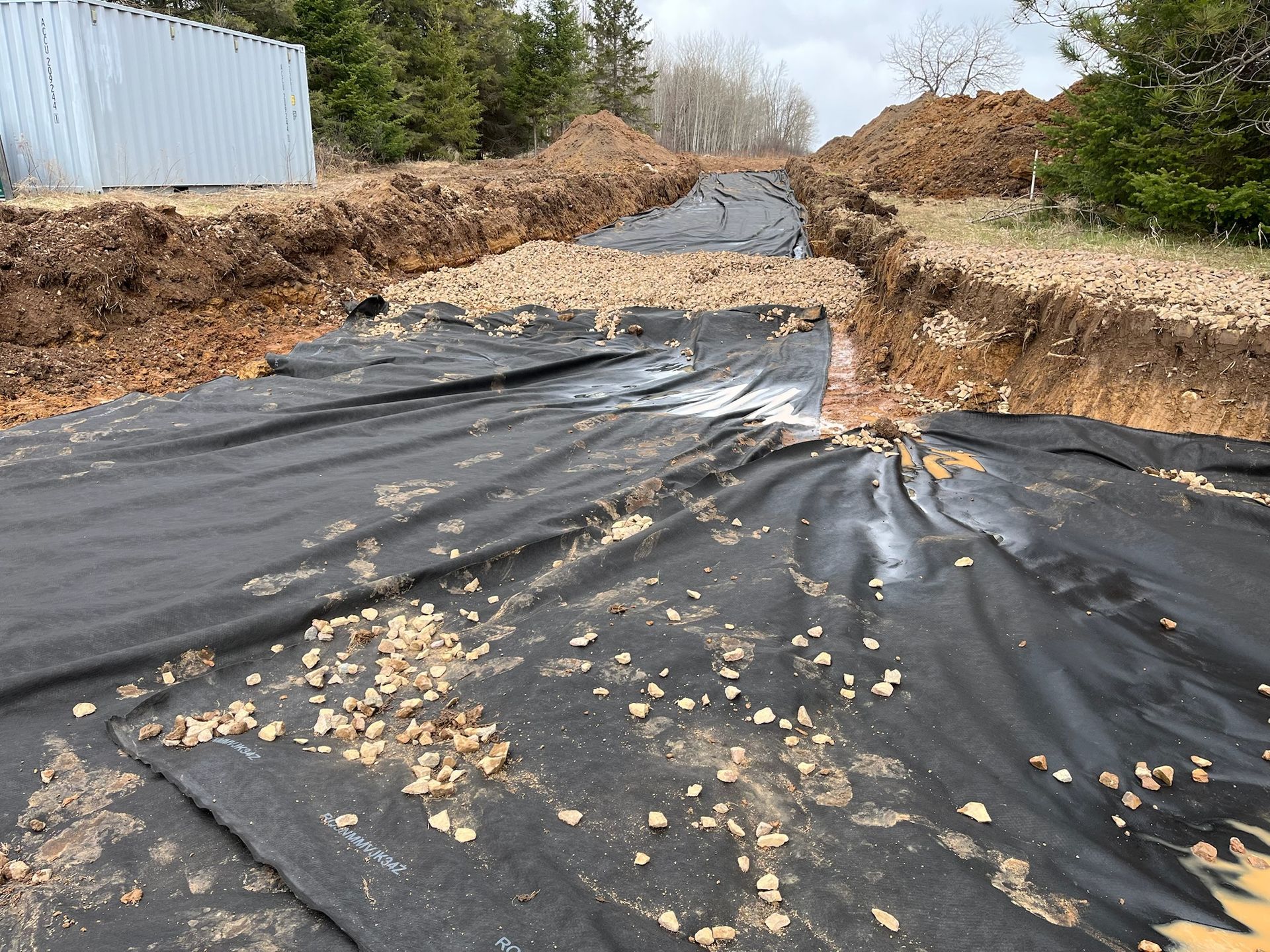 Construction site with black fabric, gravel, and a trench dug in the ground.