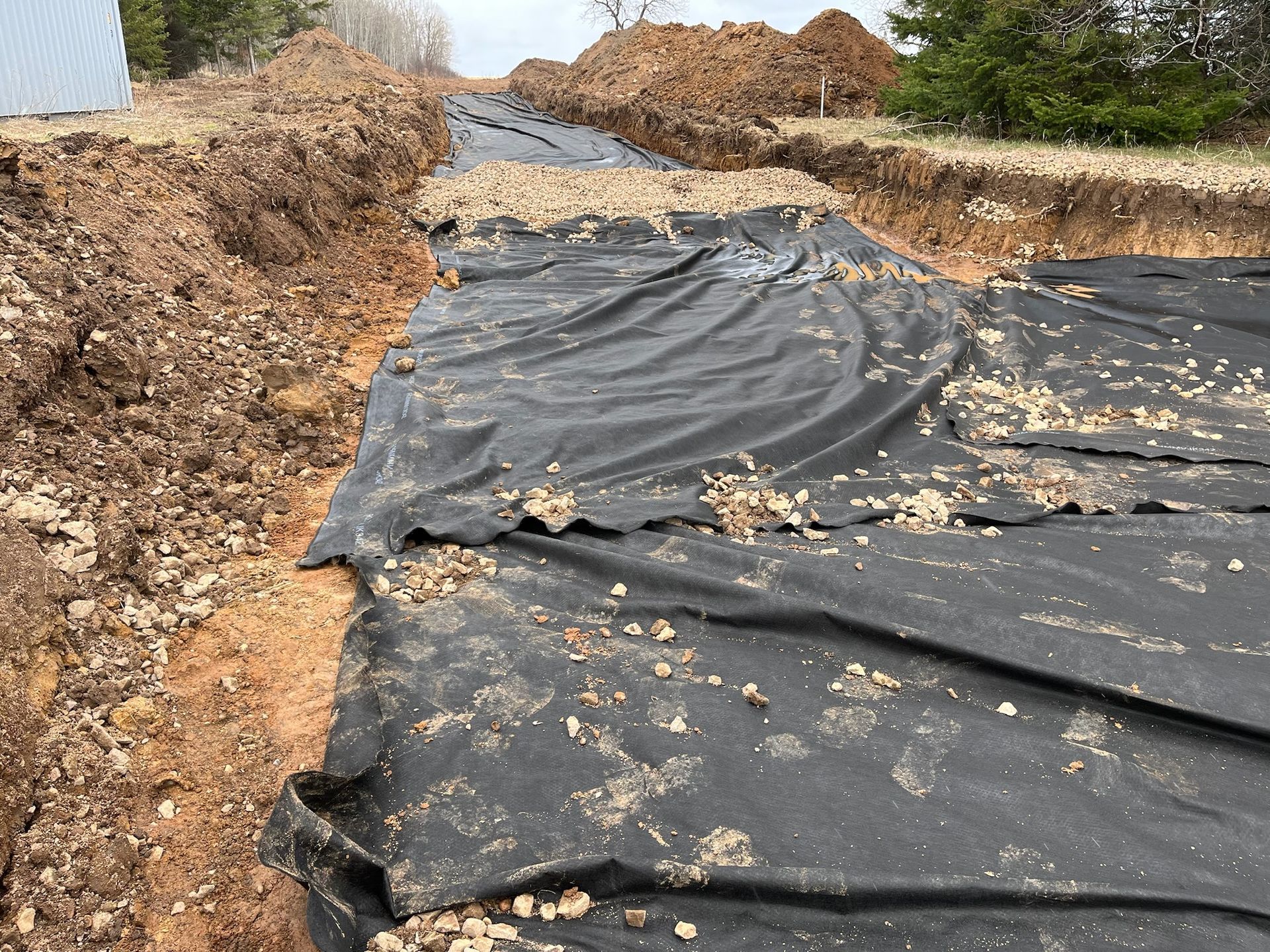 Trench excavation with black landscape fabric and gravel; dirt piles visible.