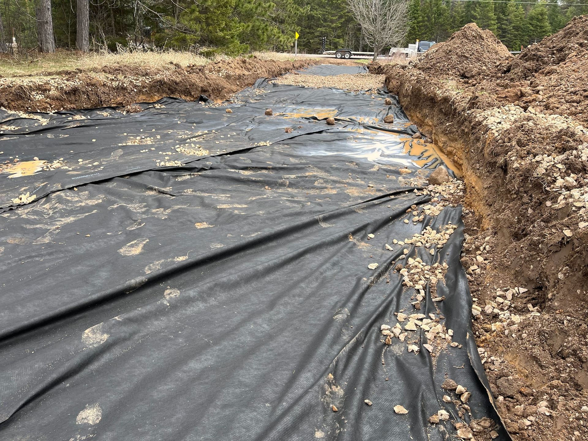 A black landscape fabric covers a trench lined with dirt. Brown soil piles border the trench.