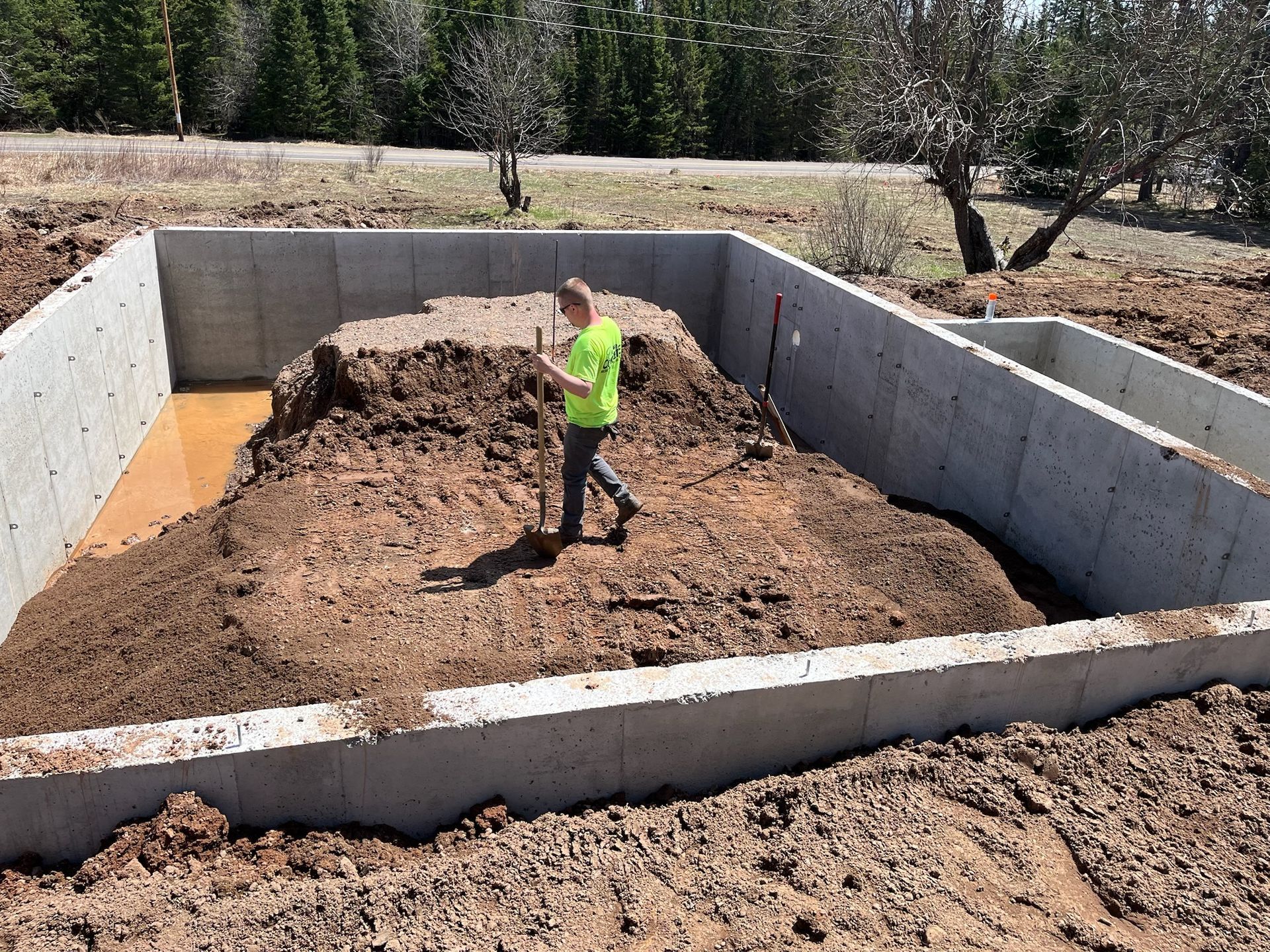 Man standing in a concrete foundation filled with soil and water. Dirt area is surrounded by concrete walls.