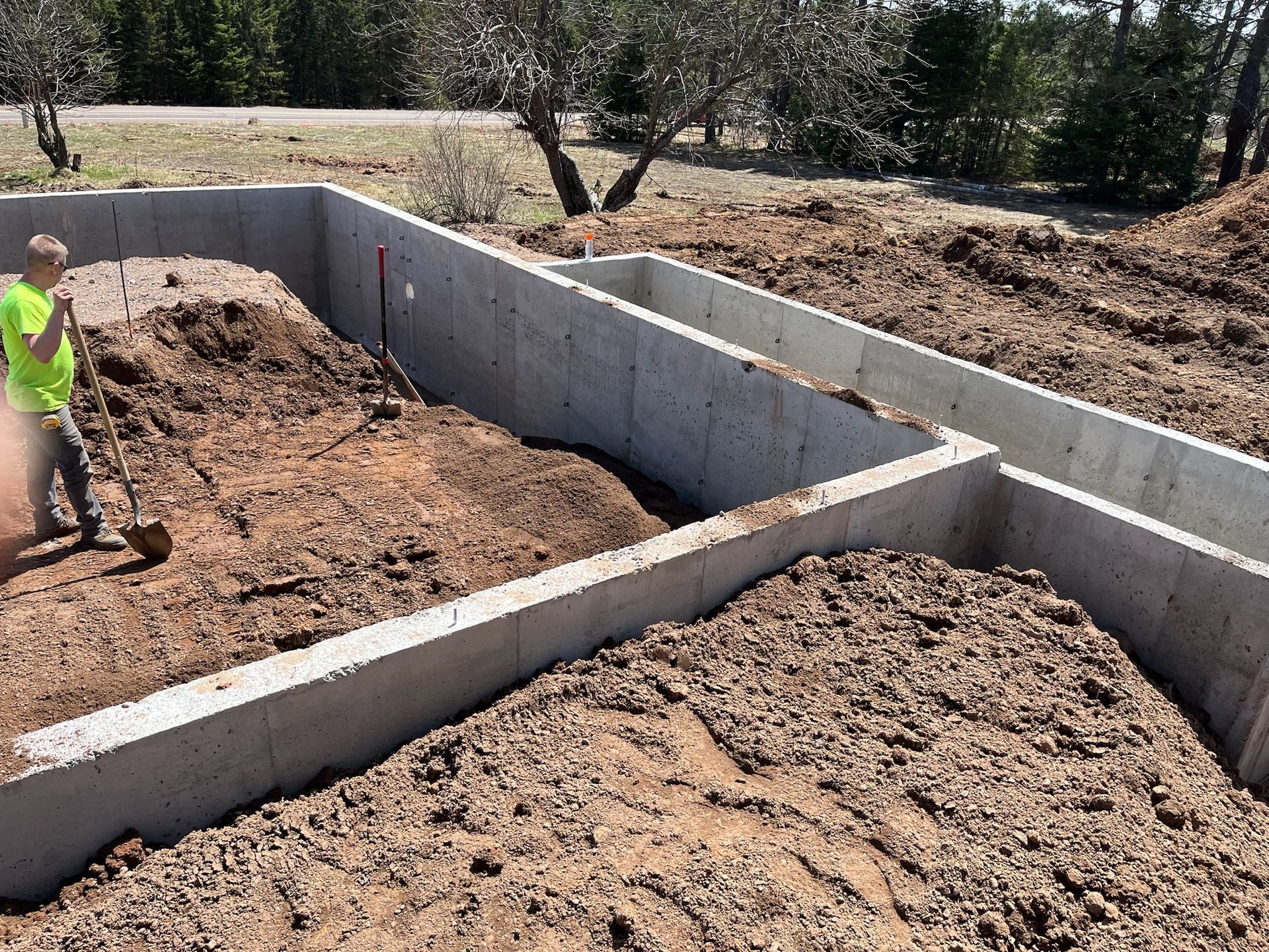 Man shoveling dirt into a concrete foundation in a yard with trees.