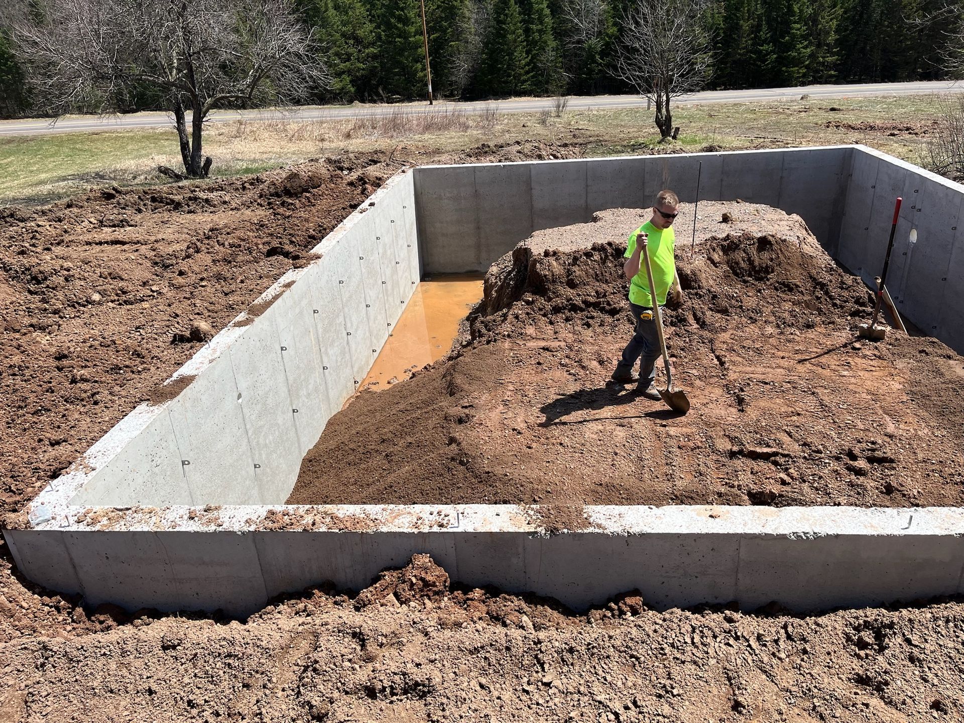 Person shoveling dirt in a concrete rectangular foundation pit, partially filled with water and soil.