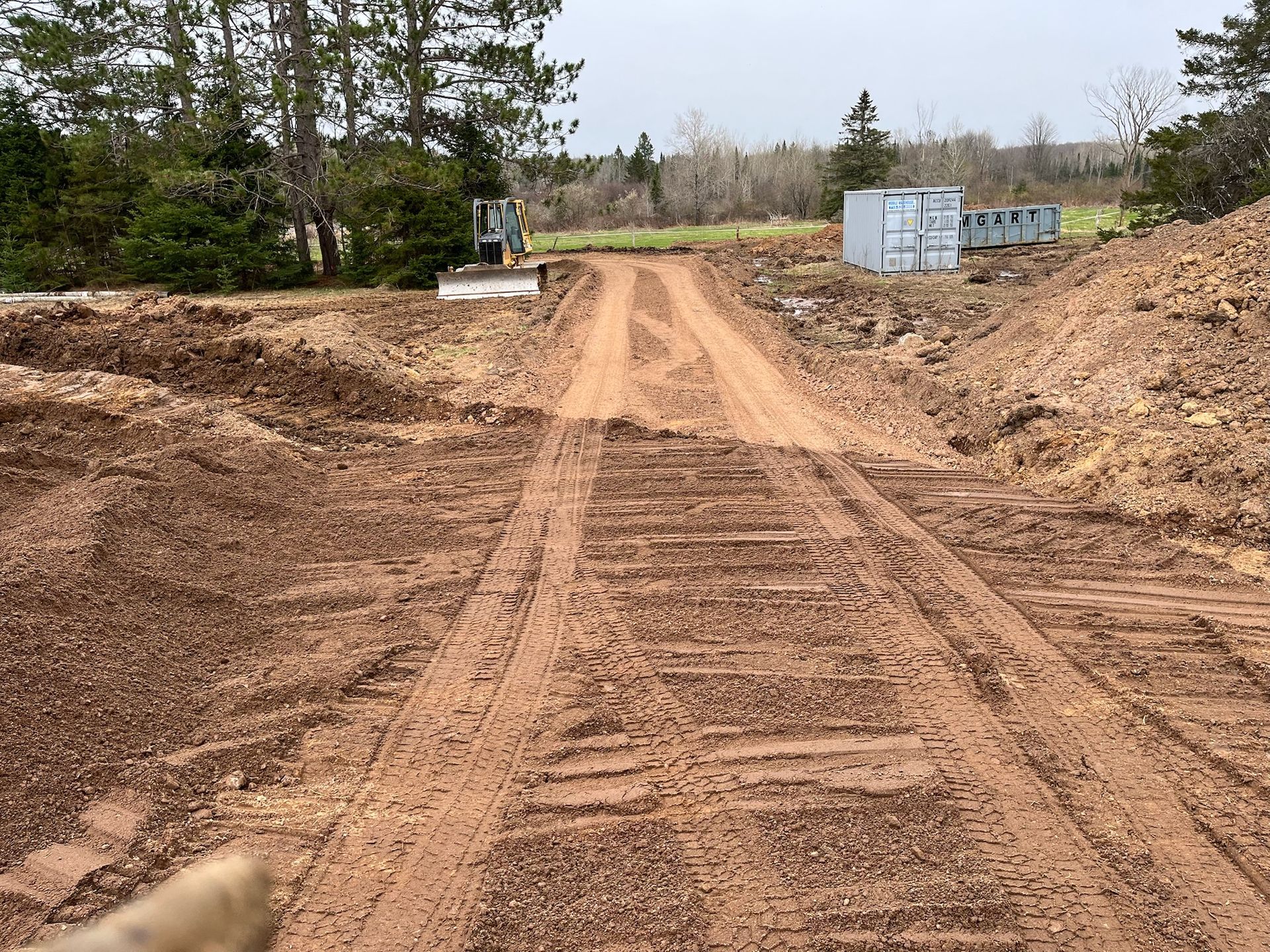 Dirt road, rutted by vehicle tracks, leads through a construction site. Trees and a storage container are in the background.