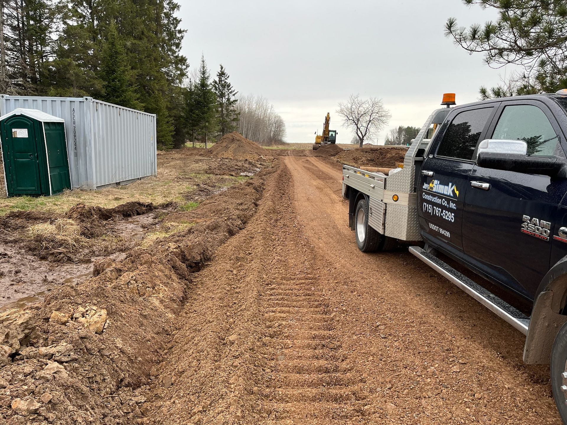 Dirt road construction site with truck and excavator, porta-potty, and overcast sky.