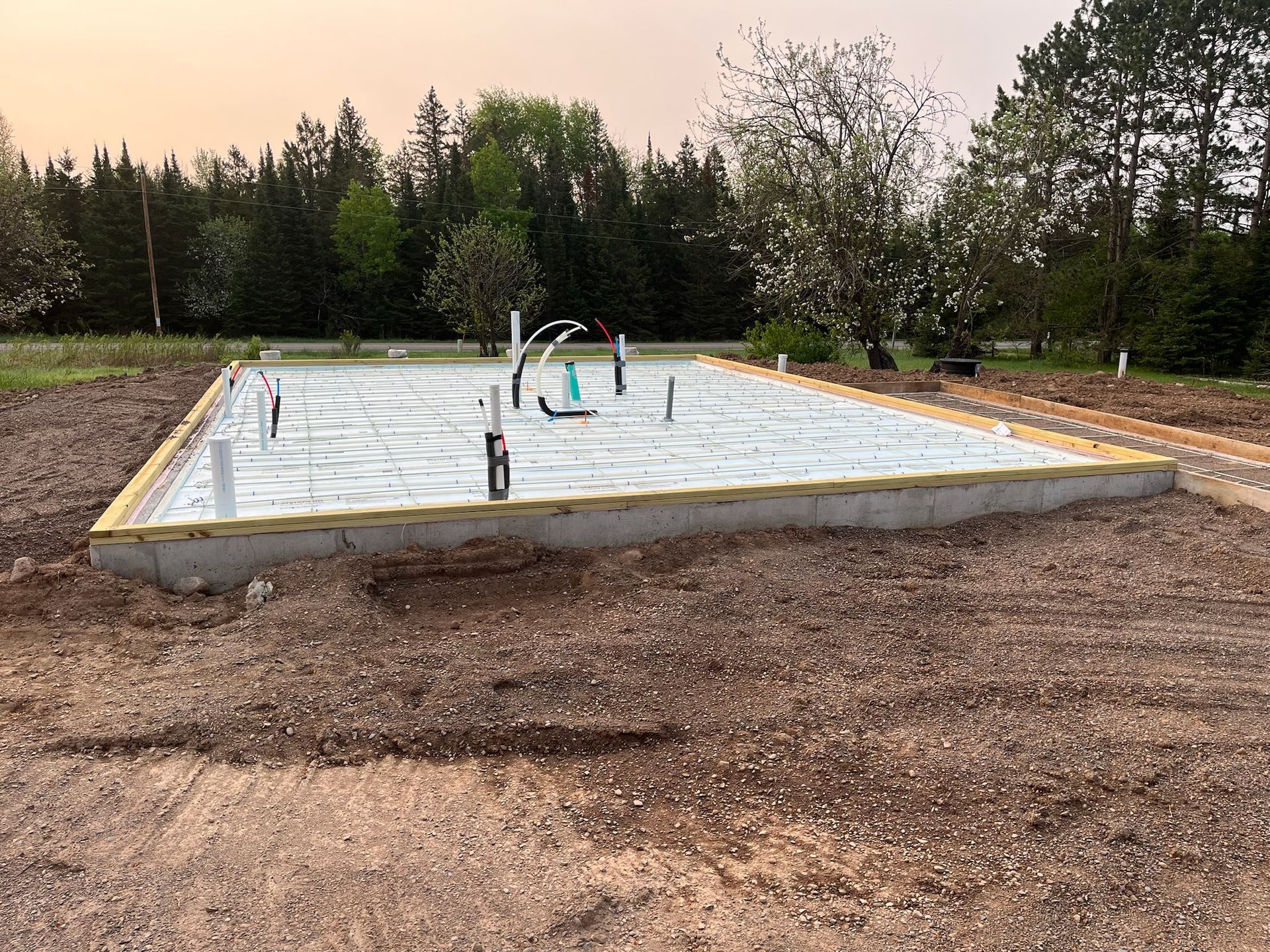 Foundation of a building, concrete slab with plumbing visible, set in a dirt lot with a treeline in the background.