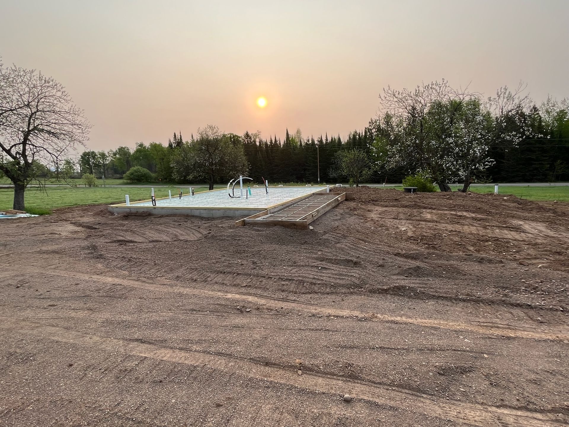 Construction site with a concrete foundation, dirt pile, and gravel. Sunset in the background.