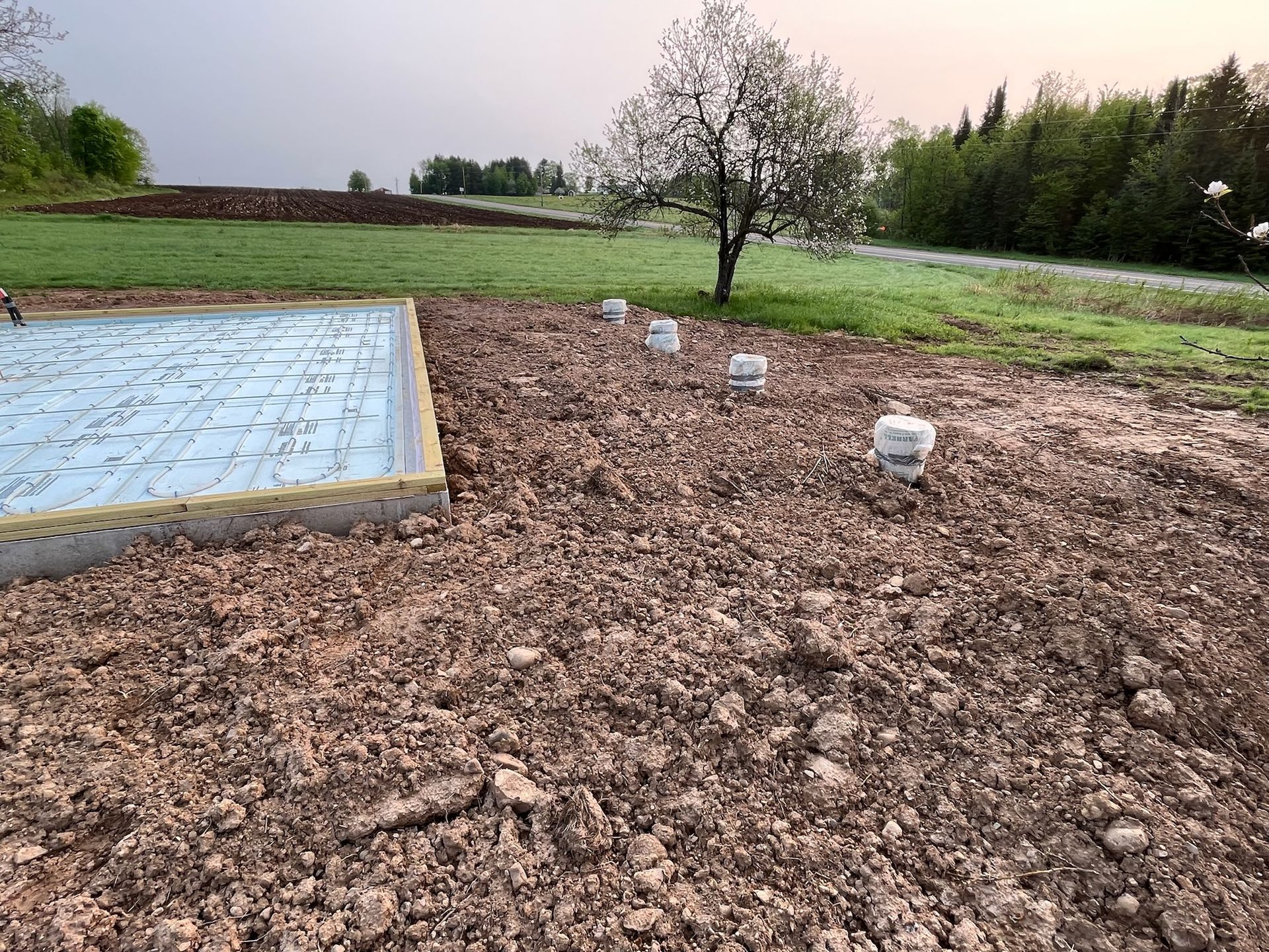 Construction site with a foundation, dirt ground, and stakes. A tree and grassy field are in the background.