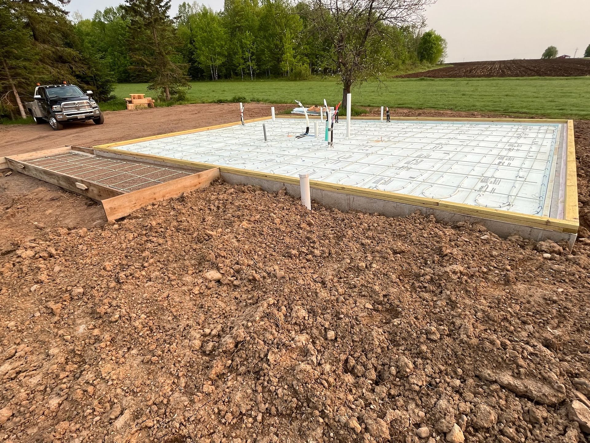 Construction site with concrete foundation and plumbing, surrounded by dirt and a truck.