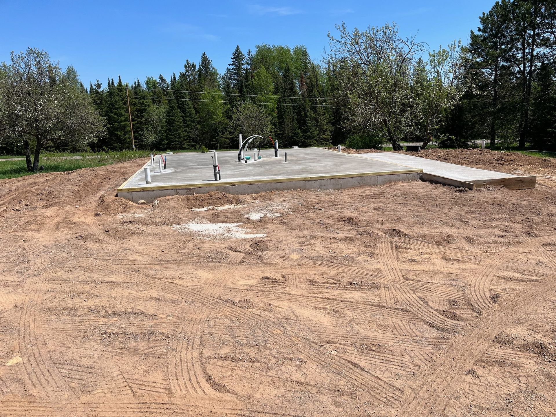 Concrete foundation with plumbing pipes on dirt lot, trees in background.