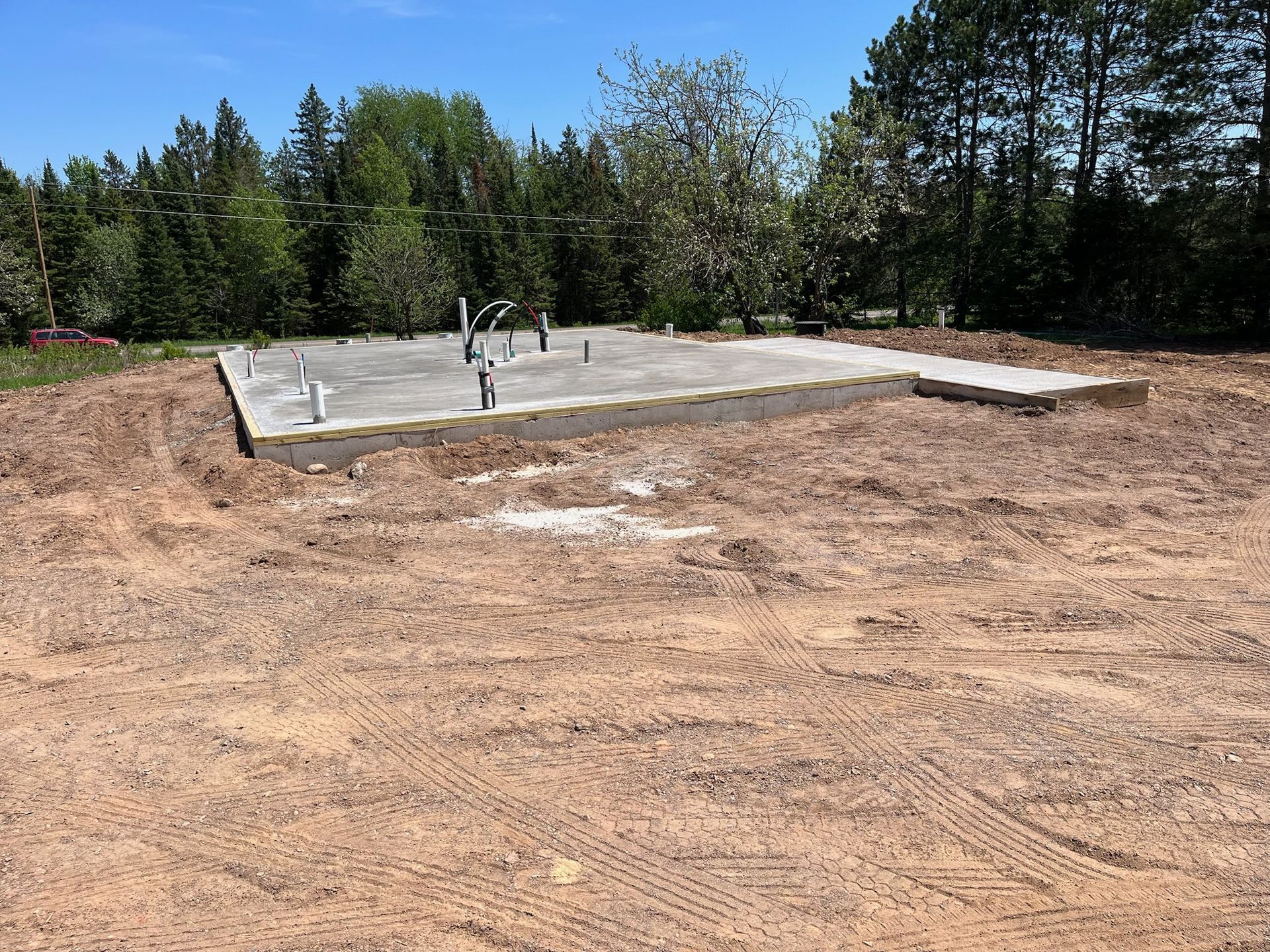 Concrete foundation with plumbing pipes on a dirt lot, trees in the background under a blue sky.