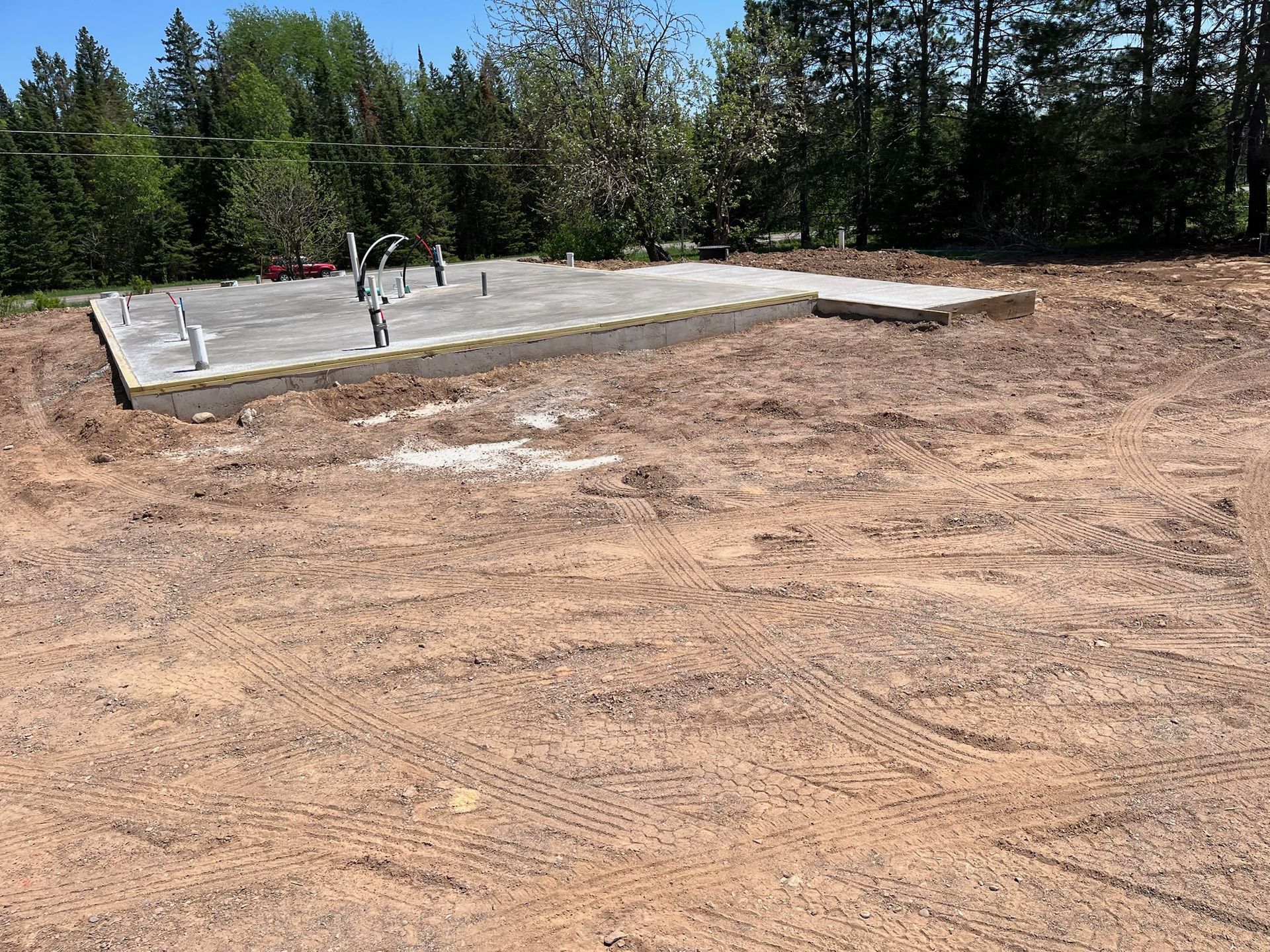 Construction site with concrete foundation and exposed plumbing, surrounded by dirt and trees.