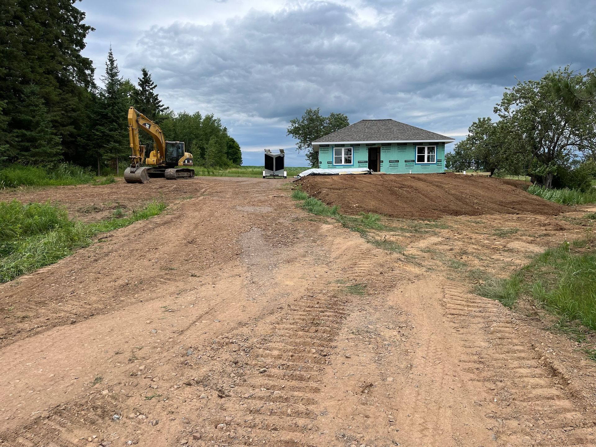 Dirt road leading to a house under construction; excavator to the left, cloudy sky.