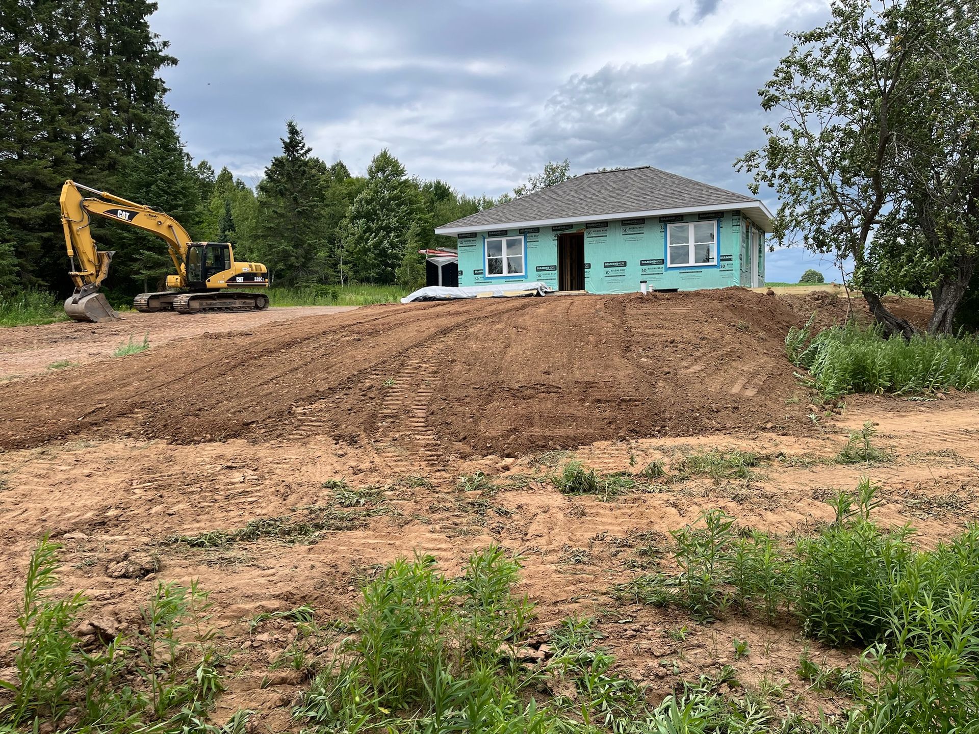 Excavator on a construction site. New house with green wrap, on a graded hill. Cloudy sky.