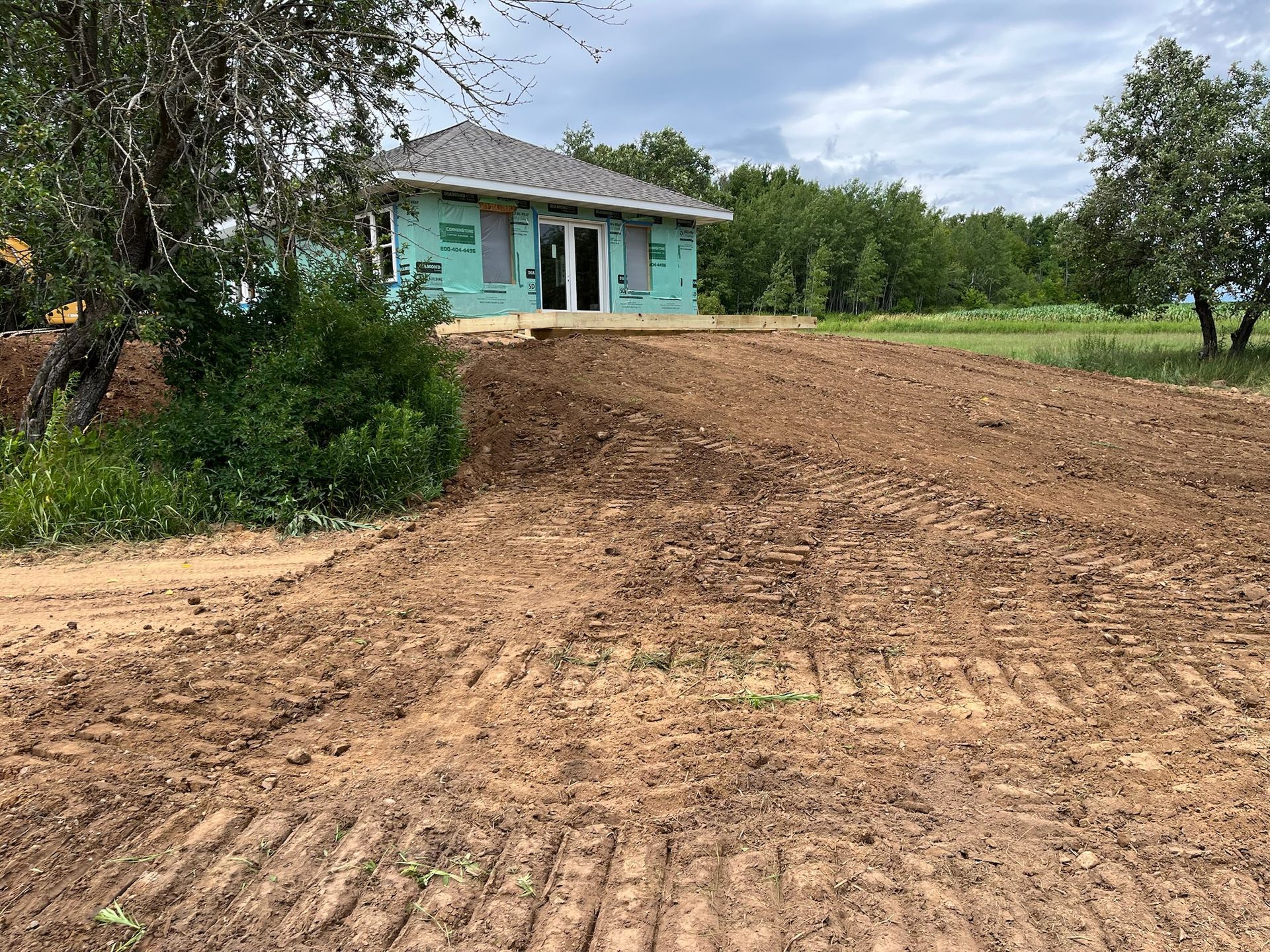 Dirt mound in front of partially built house with green wrap. Trees and field in the background.