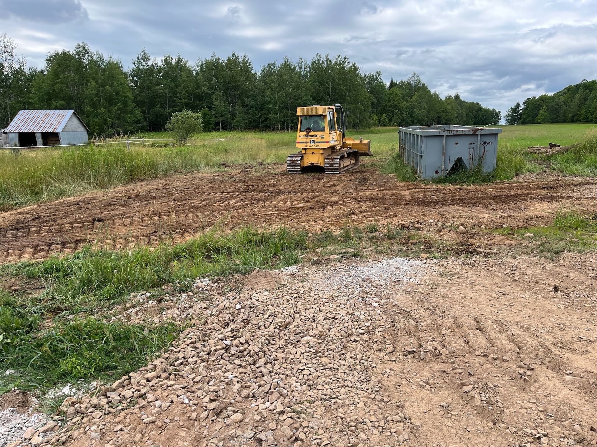 Bulldozer on a dirt path in a field, clearing land near a small building and trees.