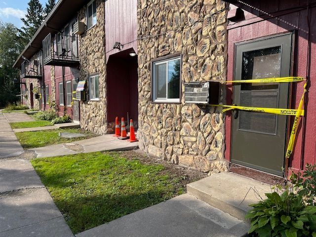 Apartment building exterior, entrance with caution tape, orange cones, stone facade, green lawn.