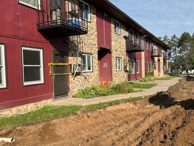 Apartment building with red siding and stone facade. Construction trench in front, caution tape at a doorway.